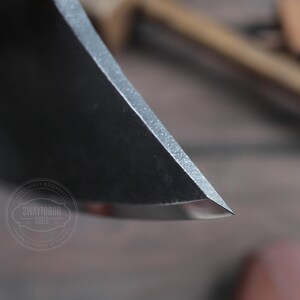 May include: Close-up of the sharp edge of a black metal axe head. The axe head is very sharp and has a slight curve to it. The axe head is in focus, while the background is blurred.