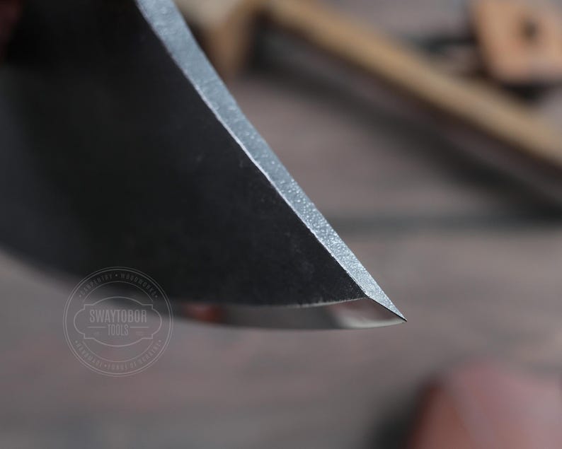 May include: Close-up of the sharp edge of a black metal axe head. The axe head is very sharp and has a slight curve to it. The axe head is in focus, while the background is blurred.