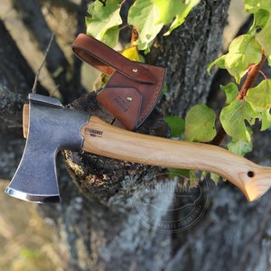Könnte beinhalten: Eine kleine, handgeschmiedete Axt mit einem Holzstiel und einer Lederscheide. Die Axt wird von Strongway Tools hergestellt, einem Unternehmen, das die Menschen in der Ukraine unterstützt.