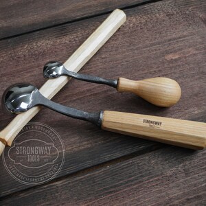 Pu&ograve; includere: Due strumenti per intagliare cucchiai con manico in legno e teste in metallo a forma di cucchiaio. Gli strumenti sono su una superficie in legno. Lo strumento pi&ugrave; grande ha il testo "Strongway Tools" sul manico.