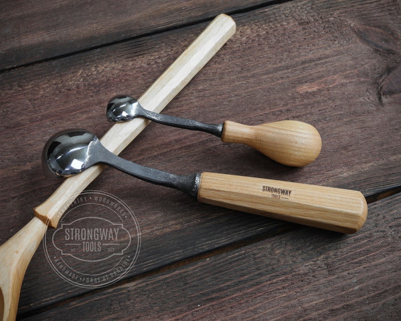 Pu&ograve; includere: Due strumenti per intagliare cucchiai con manico in legno e teste in metallo a forma di cucchiaio. Gli strumenti sono su una superficie in legno. Lo strumento pi&ugrave; grande ha il testo "Strongway Tools" sul manico.