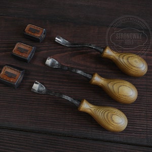 May include: Three wood carving tools with brown wooden handles and black metal blades. The tools are arranged in a row on a dark brown wooden surface. Three small leather squares with the text "Strongway Tools" are also on the surface.