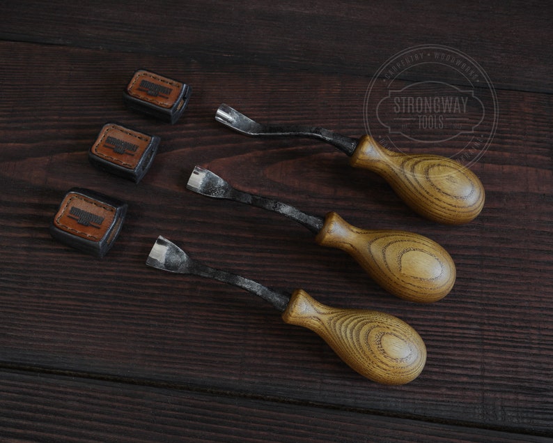 May include: Three wood carving tools with brown wooden handles and black metal blades. The tools are arranged in a row on a dark brown wooden surface. Three small leather squares with the text "Strongway Tools" are also on the surface.