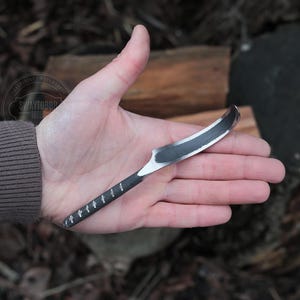 Puede incluir: Una pequeña herramienta de talla de metal negro curvada con un mango de madera. La herramienta se sostiene en una mano, con la palma hacia arriba.