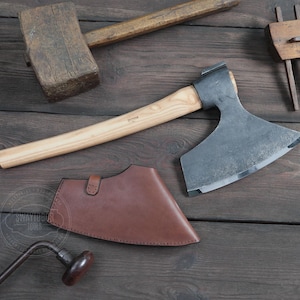 May include: A hand-forged axe with a wooden handle and a brown leather sheath. The axe has a curved blade and a rounded head. The sheath has a flap that secures with a snap.