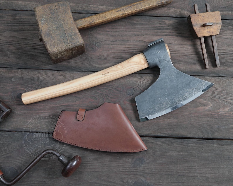 May include: A hand-forged axe with a wooden handle and a brown leather sheath. The axe has a curved blade and a rounded head. The sheath has a flap that secures with a snap.