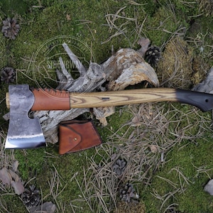 Könnte beinhalten: Eine handgeschmiedete Axt mit einer Lederschlaufe am Griff und einem Holzstiel. Die Axt hat eine schwarze Lederscheide mit einem Messingknopf. Die Axt liegt auf einem Bett aus Moos und Kiefernnadeln.