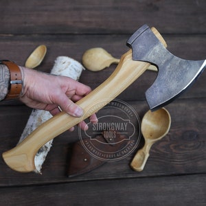 Puede incluir: Una mano sostiene un hacha hecha a mano con un mango de madera. El hacha tiene una hoja curva y un acabado oscuro. El hacha está sobre una superficie de madera con cucharas de madera y un tronco de abedul.