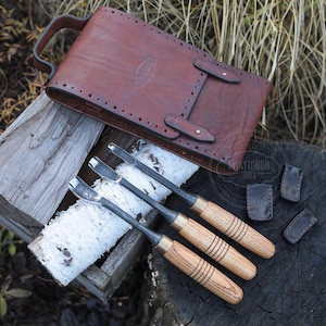Può includere: Un set di tre sgorbie per intaglio del legno con manici in legno e lame in metallo, insieme a una custodia per attrezzi in pelle marrone e tre piccoli fodere in pelle. Gli strumenti sono disposti su una superficie di legno.