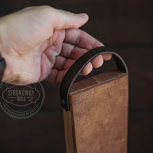 May include: A wooden cutting board with a brown leather handle. The board is made by Strongway Tools, est. 2017, and is handmade in Ukraine.