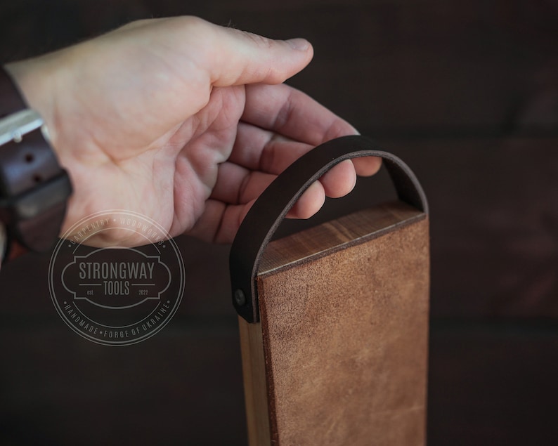 May include: A wooden cutting board with a brown leather handle. The board is made by Strongway Tools, est. 2017, and is handmade in Ukraine.