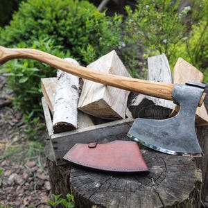 Può includere: Ascia con manico in legno e testa in metallo, appoggiata su un ceppo di legno. Un fodero in pelle marrone è accanto all'ascia. L'ascia è accanto a una cassa di legno piena di legna da ardere. L'ascia ha il testo "SWAYTOBO TOOLS".
