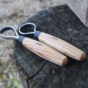 May include: Two wood carving tools with metal loop handles rest on a weathered wooden surface. The handles are light-colored wood with a dark gray band near the metal loop. The tools appear to be new.