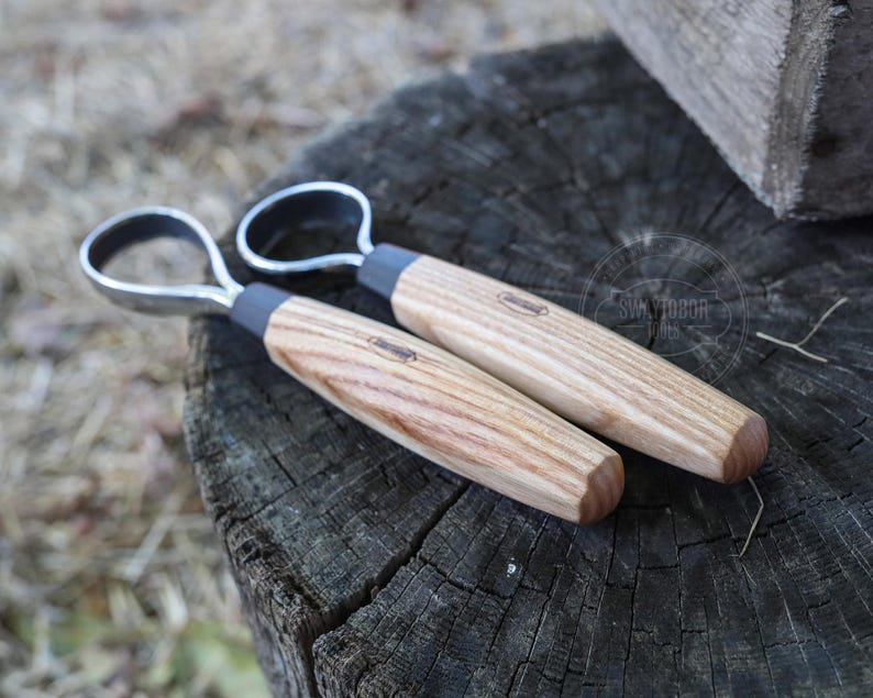 May include: Two wood carving tools with metal loop handles rest on a weathered wooden surface. The handles are light-colored wood with a dark gray band near the metal loop. The tools appear to be new.