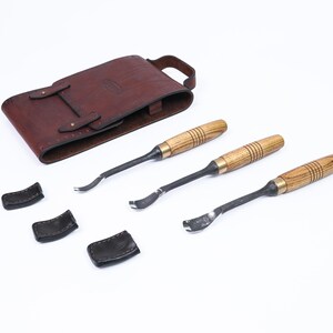 Może przedstawiać: Zestaw trzech narzędzi do rzeźbienia w drewnie z drewnianymi uchwytami i metalowymi ostrzami, wraz z brązowym sk&oacute;rzanym etui i trzema czarnymi sk&oacute;rzanymi osłonami ostrzy. Narzędzia są ułożone na białej powierzchni.