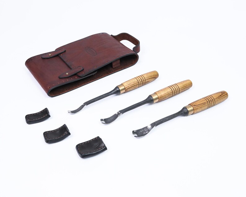 Może przedstawiać: Zestaw trzech narzędzi do rzeźbienia w drewnie z drewnianymi uchwytami i metalowymi ostrzami, wraz z brązowym sk&oacute;rzanym etui i trzema czarnymi sk&oacute;rzanymi osłonami ostrzy. Narzędzia są ułożone na białej powierzchni.
