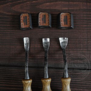 May include: Three wood carving tools with brown wooden handles and metal blades. The tools are arranged in a row on a dark brown wooden surface. The tools are labeled "Strongway Tools" in a white circle.