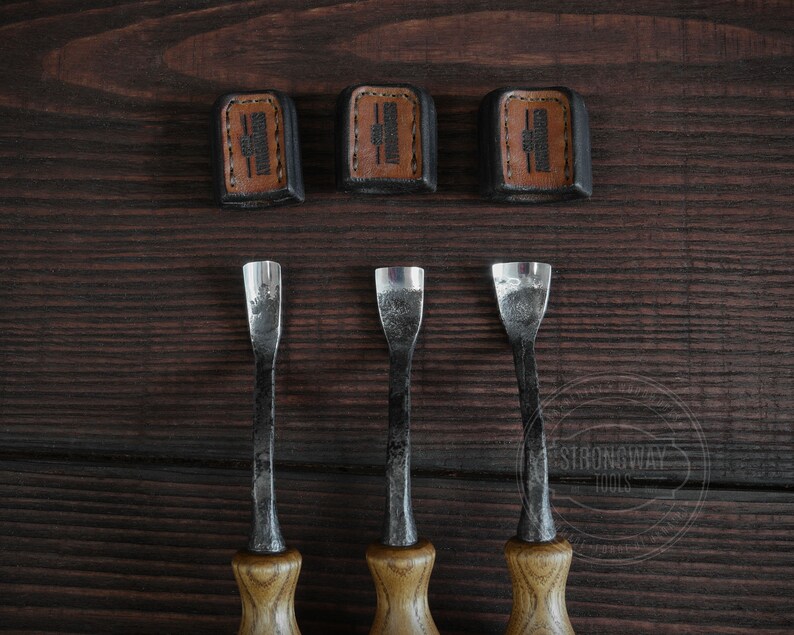 May include: Three wood carving tools with brown wooden handles and metal blades. The tools are arranged in a row on a dark brown wooden surface. The tools are labeled "Strongway Tools" in a white circle.