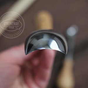 Pu&ograve; includere: Primo piano di uno strumento da intaglio in metallo curvo, lucido e color argento. Lo strumento &egrave; tenuto in una mano, con lo sfondo sfocato.