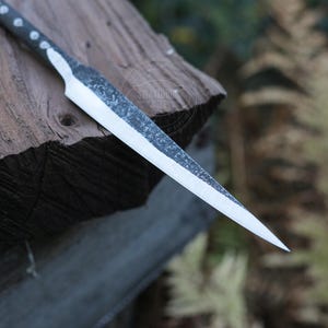 May include: A handcrafted, sharp-edged knife with a textured blade and a dark handle. The knife is resting on a piece of weathered wood, showcasing its rustic design and craftsmanship. The blade is approximately 8 inches long.
