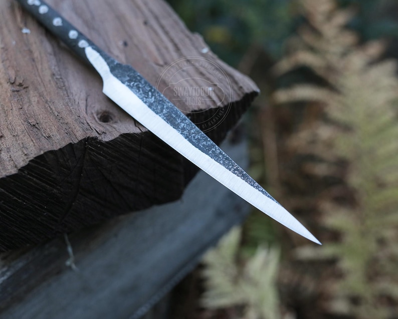 May include: A handcrafted, sharp-edged knife with a textured blade and a dark handle. The knife is resting on a piece of weathered wood, showcasing its rustic design and craftsmanship. The blade is approximately 8 inches long.
