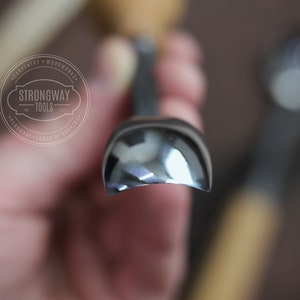 Pu&ograve; includere: Primo piano di uno strumento per la lavorazione del legno curvo, lucido e argentato. Lo strumento &egrave; tenuto in una mano e lo sfondo &egrave; sfocato. Lo strumento &egrave; etichettato "Strongway Tools" con il testo "Carpentry - Woodworks - Handmade - Forge de Ukraine".