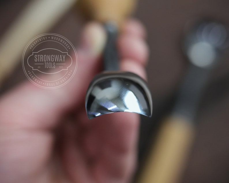 Pu&ograve; includere: Primo piano di uno strumento per la lavorazione del legno curvo, lucido e argentato. Lo strumento &egrave; tenuto in una mano e lo sfondo &egrave; sfocato. Lo strumento &egrave; etichettato "Strongway Tools" con il testo "Carpentry - Woodworks - Handmade - Forge de Ukraine".