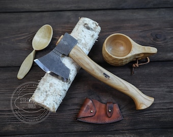 May include: A handcrafted wooden axe with a leather sheath, a wooden spoon and a wooden cup on a birch log background. The axe has a black metal head and a wooden handle. The text "Strong Tools" is visible on the log.