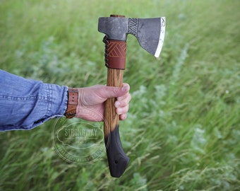 May include: A handcrafted axe with a wooden handle and a metal blade. The handle is stained dark at the bottom and features a brown leather wrap. The axe is held against a backdrop of green grass.