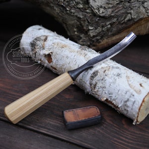 May include: A curved wood carving knife with a wooden handle. The knife is resting on a piece of birch wood. The knife is made by Strongway Tools, Handmade Forge of Ukraine.