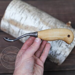 May include: A small, curved wood carving tool with a wooden handle and a metal blade. The tool is held in a hand against a birch bark background.