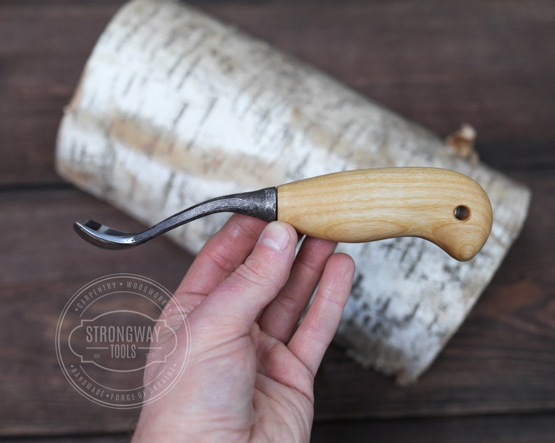 May include: A small, curved wood carving tool with a wooden handle and a metal blade. The tool is held in a hand against a birch bark background.