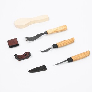 Può includere: Una collezione di attrezzi per intaglio del legno, tra cui un grezzo di cucchiaio, tre coltelli con manici in legno e due foderi in pelle. Le lame sono argentate e i manici sono in legno chiaro. I foderi in pelle sono marroni e neri.