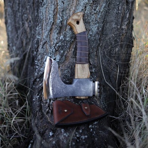 Op de afbeelding: Een handgesmeed bijl met een houten steel en een leren schede. De bijl leunt tegen een boomstam met een logo voor "Strongwood Tools" zichtbaar op de achtergrond.