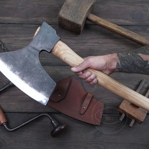 May include: A hand holding a large, curved axe with a wooden handle. The axe has a dark gray blade and a light brown handle. The axe is resting on a wooden surface with a leather sheath and other tools.