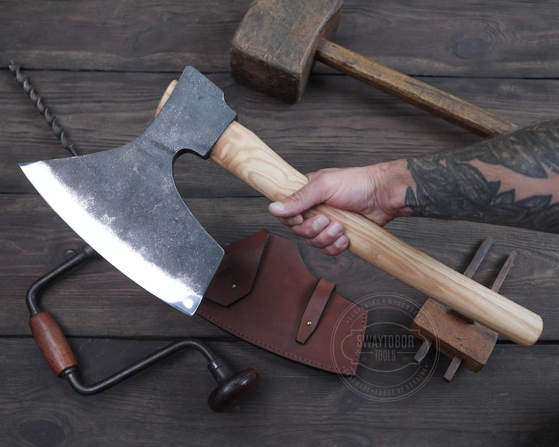 May include: A hand holding a large, curved axe with a wooden handle. The axe has a dark gray blade and a light brown handle. The axe is resting on a wooden surface with a leather sheath and other tools.