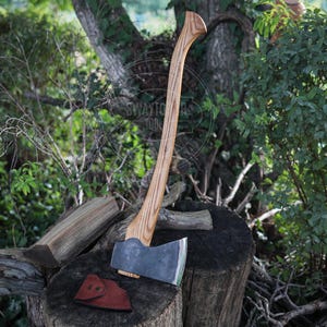 Può includere: Un'ascia da abbattimento con testa in metallo scuro e un lungo manico in legno curvo con un motivo a venature distinto. Un piccolo fodero in pelle marrone con bottone a pressione poggia accanto alla testa dell'ascia, marcata con il testo SWAYTOOK TOOLS.