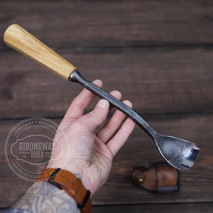 May include: A hand holds a wooden handled spoon carving tool with a metal scoop. The tool is made by Strongway Tools and is handmade and forged.