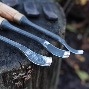 Może przedstawiać: Zbliżenie trzech dłut do rzeźbienia w drewnie z drewnianymi uchwytami i metalowymi ostrzami. Ostrza mają zakrzywiony kształt. Uchwyty są jasnobrązowe z mosiężną tuleją. Narzędzia spoczywają na zwietrzałej drewnianej powierzchni.