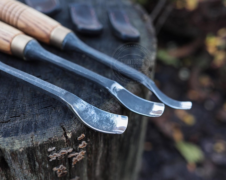 Może przedstawiać: Zbliżenie trzech dłut do rzeźbienia w drewnie z drewnianymi uchwytami i metalowymi ostrzami. Ostrza mają zakrzywiony kształt. Uchwyty są jasnobrązowe z mosiężną tuleją. Narzędzia spoczywają na zwietrzałej drewnianej powierzchni.