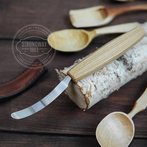 以下が含まれることがあります： 木製のハンドルが付いた湾曲した木彫りナイフ。ナイフは、木のさじが隣に置かれた白樺の丸太の上に置かれています。ナイフは、ウクライナで手作り工具を製造するStrongway Toolsによって作られています。