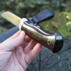 May include: A handcrafted knife with a polished, gold-toned blade and a dark wooden handle. The handle has a curved design with a black end cap and a small, decorative brass accent. The knife is designed for outdoor use.
