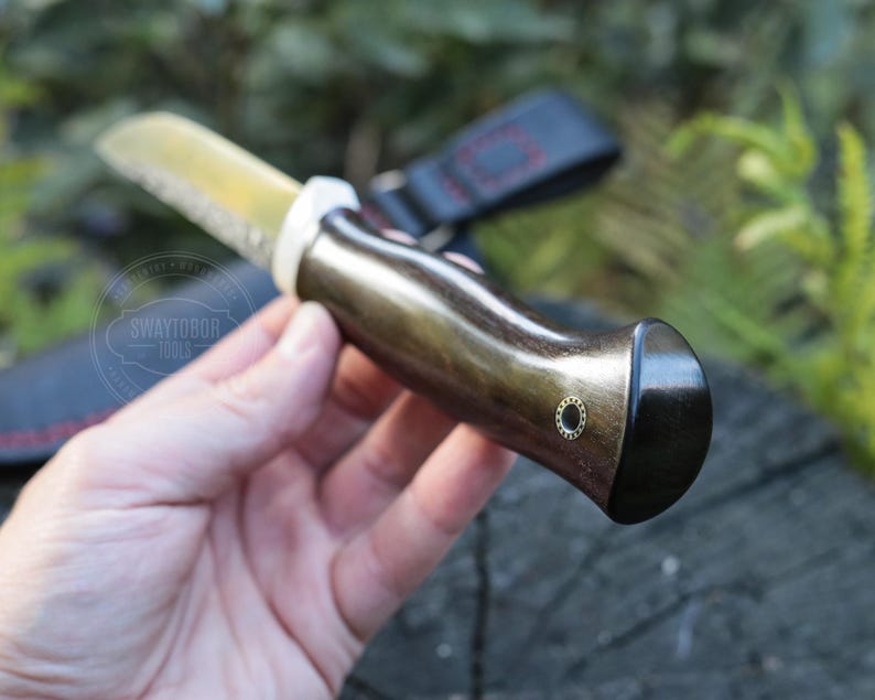 May include: A handcrafted knife with a polished, gold-toned blade and a dark wooden handle. The handle has a curved design with a black end cap and a small, decorative brass accent. The knife is designed for outdoor use.