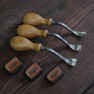 May include: Three wood handled carving tools with curved blades. The tools are arranged in a row on a dark wood surface. Each tool has a leather label with the text "Strongway Tools" on it.