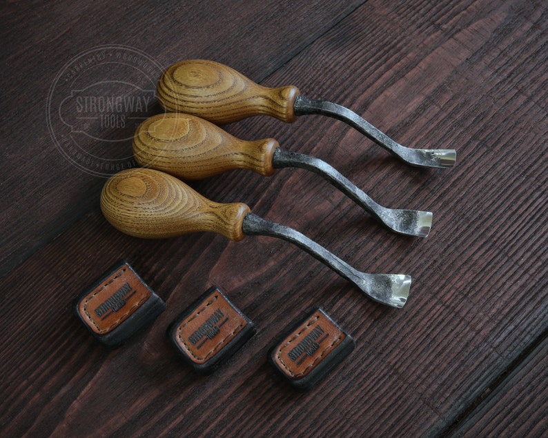 May include: Three wood handled carving tools with curved blades. The tools are arranged in a row on a dark wood surface. Each tool has a leather label with the text "Strongway Tools" on it.