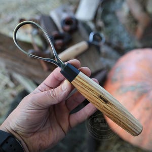 May include: A wood carving hook knife with a curved blade and a wooden handle. The handle has a black ferrule and a light brown wood grain pattern. The knife is held in a hand, with a pumpkin in the background.