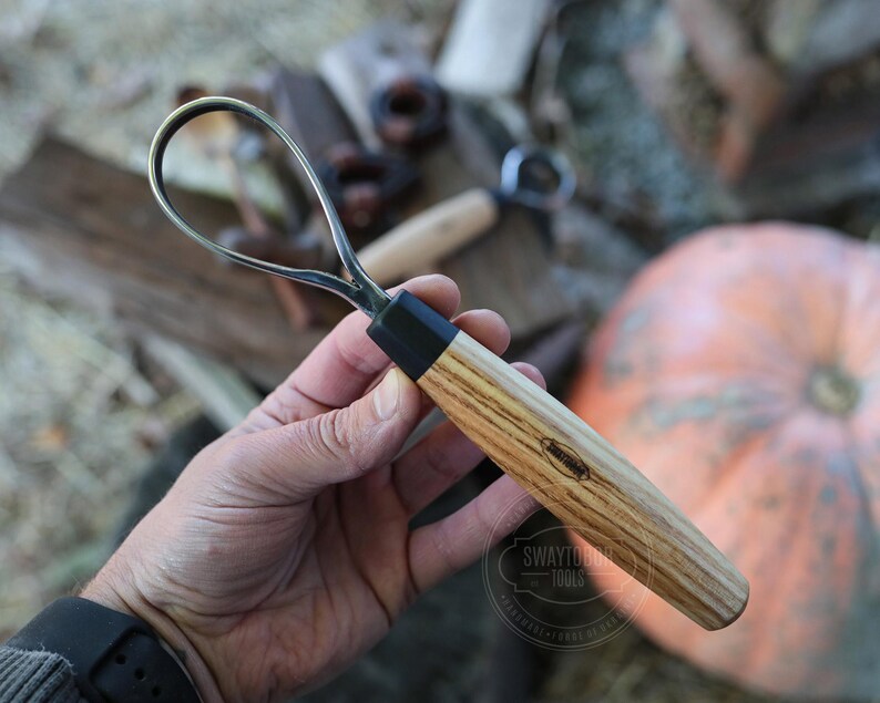 May include: A wood carving hook knife with a curved blade and a wooden handle. The handle has a black ferrule and a light brown wood grain pattern. The knife is held in a hand, with a pumpkin in the background.