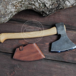 May include: A hand-forged axe with a wooden handle and a leather sheath. The axe head is made of steel and has a curved blade. The handle is made of light wood and has a slight curve. The sheath is made of brown leather and has a stitched design.