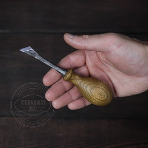 Puede incluir: Un formón para carpintería con mango de madera y hoja de metal. El formón se sostiene en una mano, mostrando el diseño y la artesanía de la herramienta. El mango es de color marrón claro y la hoja es plateada.