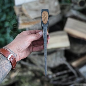 May include: A hand-forged steel axe head featuring a dark, textured metal finish and a light-colored, oval-shaped wooden inlay on the top edge. The tool displays a sharp, tapered profile and includes the etched text SWAYTOBOR TOOLS on the side.